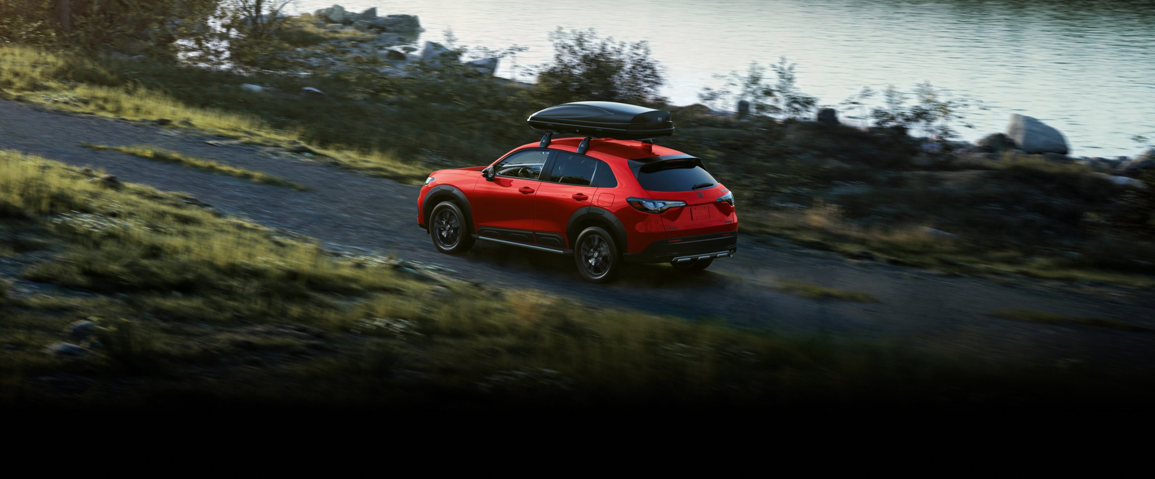2027 Honda HR-V Sport in Milano Red profile view driving on a dirt road near water with a roof box (Subcompact Crossover SUV).