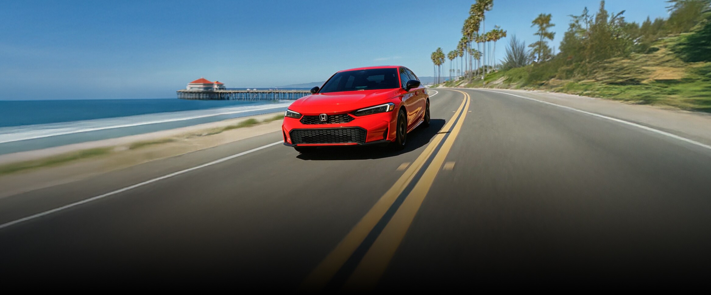 2026 Honda Civic Sedan Sport Hybrid in Rallye Red front driving on a scenic coastal road by the ocean (Compact Sedan).