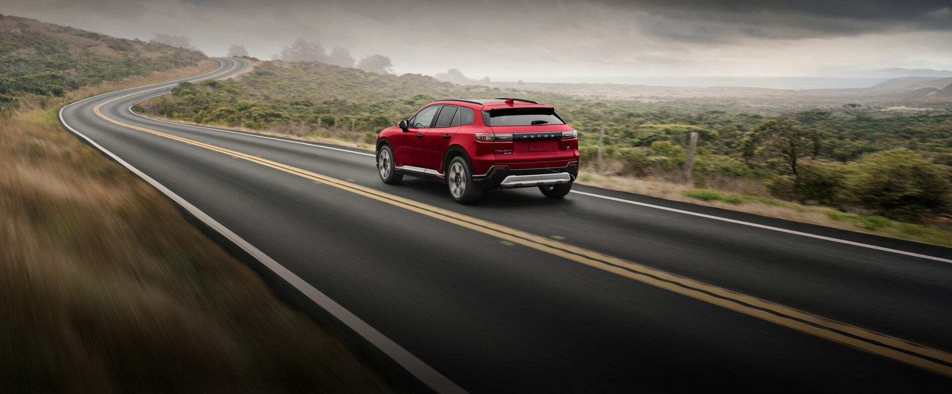 2026 Honda Prologue Elite in Scarlet Red Pearl, driving on a coastal road on a moody day.