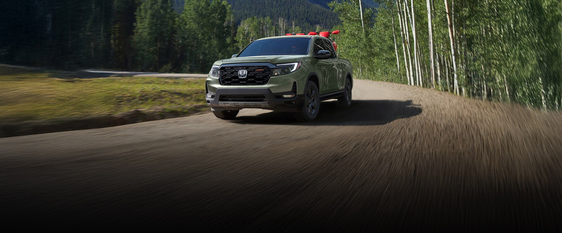 2026 Ridgeline TrailSport in Modern Steel Metallic drives through a dirt field, carrying 2 dirt bikes in the flat bed and towing a Honda Talon on a trailer.