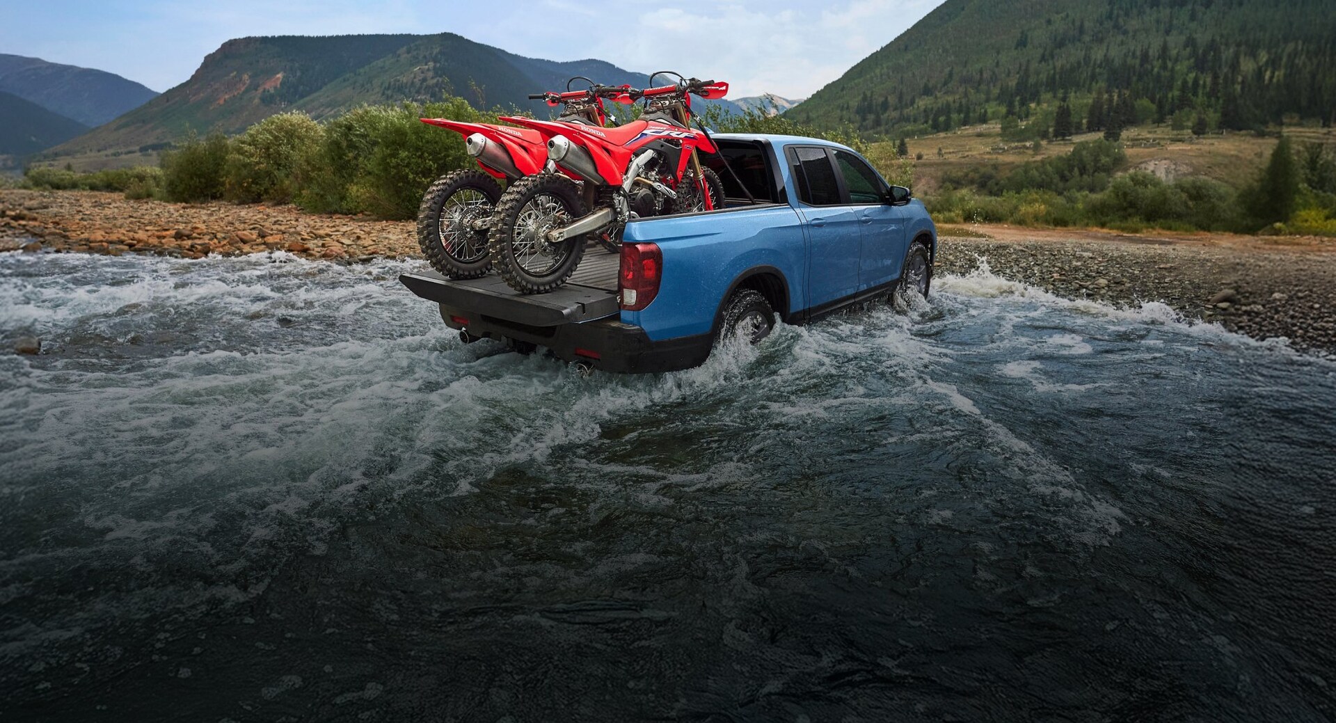 2025 Honda Ridgeline TrailSport in Diffused Sky Blue Pearl carrying 2x CRF motorbikes in the tuck bed while driving across a small river in the mountains in Colorado.