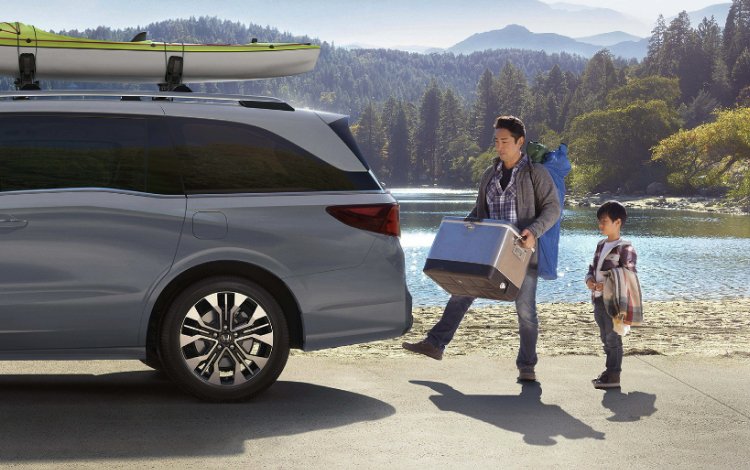 2026 Honda Odyssey Elite parked at lake with hills and greenery in background. Father  using the motion sensor to open the back door to load items while son is watching.