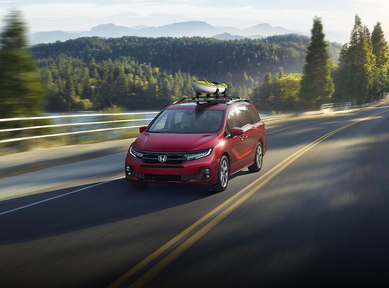 2026 Honda Odyssey Elite in Radiant Red Metallic II driving on a paved country road, view of greenery and rolling hills in background. 2026 Honda Odyssey Elite in Radiant Red Metallic II driving on a paved country road, view of greenery and rolling hills in background.