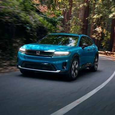 2026 Honda Prologue Elite in North Shore Pearl, driving through redwoods, taking a curved turn on a paved road, with light peeking through the trees in the background.