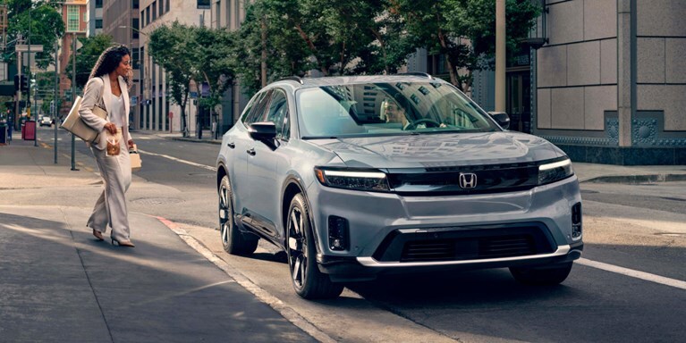 2026 Honda Prologue Elite in Sonic Gray Pearl, parked curbside at an urban café, with a business woman approaching the passenger side of the vehicle to greet a man in the driver’s seat.