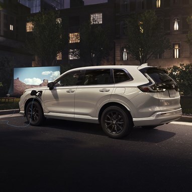 Driver's side rear view of 2026 CR-V e:FCEV in Platinum White Pearl in a downtown loft parking lot showing an outdoor movie night on the side of a building, with the projector plugged into the exterior power charger.