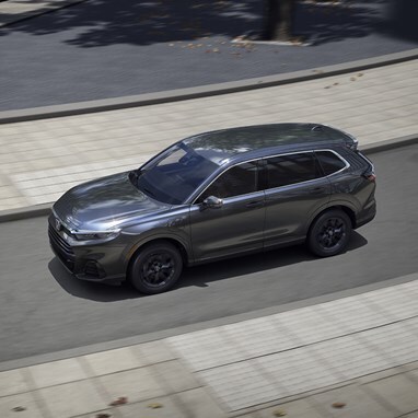 High driver profile view of 2026 CR-V e:FCEV in Meteorite Gray Metallic driving on a curved road in a downtown area.