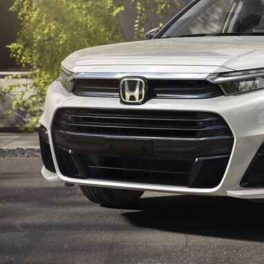 Grille and LED Headlight detail of 2026 CR-V e:FCEV in Platinum White Pearl parked in a clean modern urban location, with trees / greenery.
