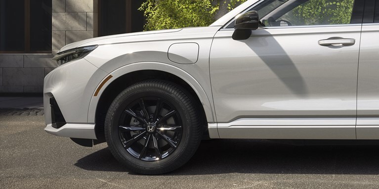 Wheel detail of 2026 CR-V e:FCEV in Platinum White Pearl showing 18" alloy wheels parked in a clean modern urban location, with trees / greenery.