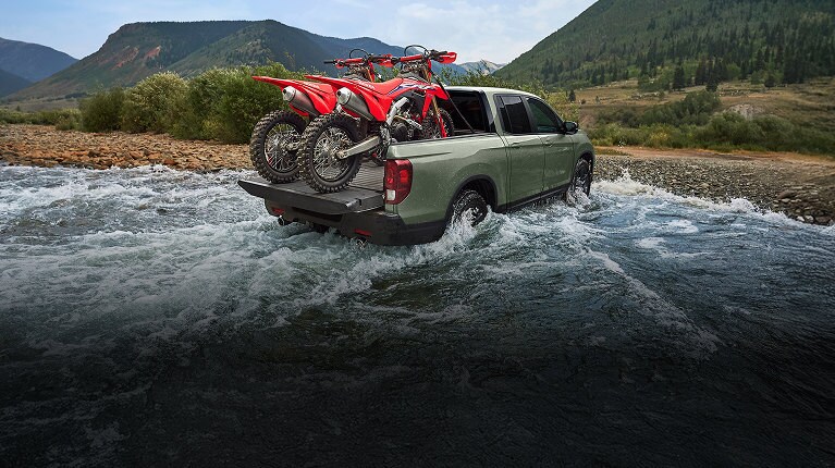 2026 Honda Ridgeline TrailSport in Ash Green Metallic carrying 2 motorbikes in the tuck bed while driving across a small river in the mountains 2026 Honda Ridgeline TrailSport in Ash Green Metallic carrying 2 motorbikes in the tuck bed while driving across a small river in the mountains
