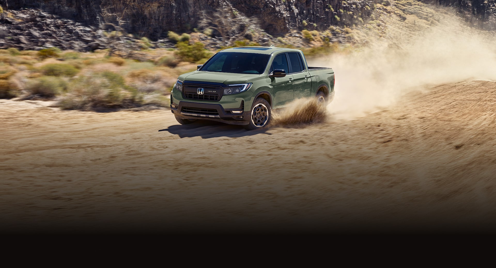 2026 Ridgeline TrailSport in Ash Green Metallic driving  through rugged terrain with dust kicking up,  with mountains in background, at sunrise.