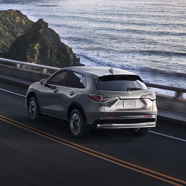 2027 Honda HR-V EX-L in Modern Steel Metallic rear view driving on a coastal road near the ocean (Subcompact Crossover SUV).