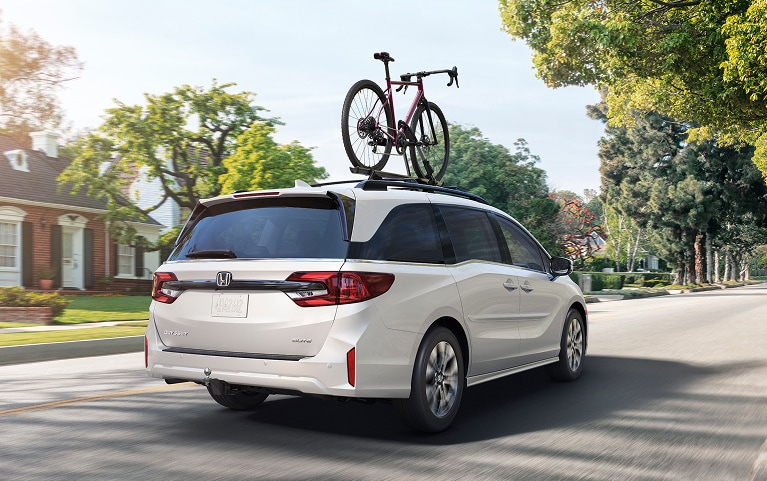 2027 Honda Odyssey Elite in Platinum White Pearl back driving through a sunny residential neighborhood (Minivan).