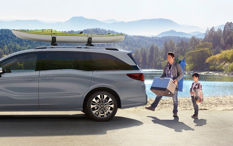 2027 Honda Odyssey Elite in Sonic Gray Pearl profile parked by a scenic lake with a family loading gear (Minivan).