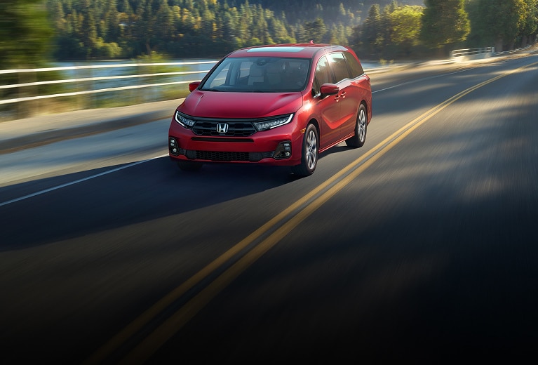 2027 Honda Odyssey Elite in Radiant Red Metallic II front driving on a scenic road near a lake and forest (Minivan).